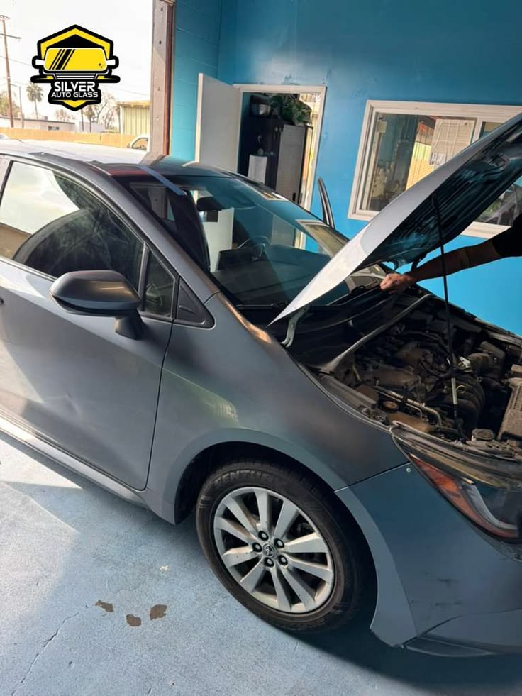 Silver sedan with open hood at auto glass shop with blue building in background